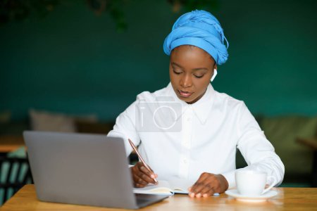 Photo for Cheerful beautiuful young black woman in smart casual entrepreneur attending online business meeting, lady sit at table in front of laptop, using earpods, taking notes, cafe interior, copy space - Royalty Free Image