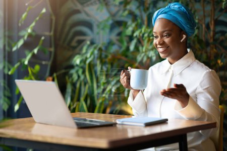 Photo for Positive beautiful millennial black lady entrepreneur have online meeting, sitting at table in front of laptop, using earpods, drinking cofee, businesswoman working at cafe, copy space - Royalty Free Image