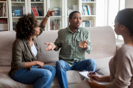 Téléchargez les photos : Emotional young black couple fighting in front of family counselor, furious angry unhappy african husband and wife sitting on couch at psychologist office, yelling and gesturing. Marriage counseling - en image libre de droit