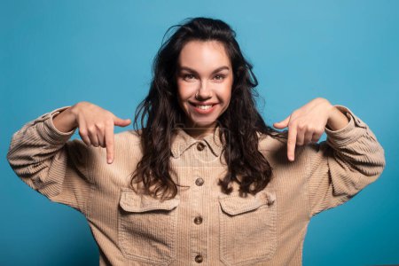 Foto de Happy millennial european female brunette student showing thumbs down to empty space, isolated on blue background, studio. Sale recommendation and advice, lifestyle, ad and offer, facial expression - Imagen libre de derechos