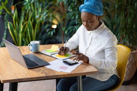 Pretty young black businesswoman in smart casual and head turban working at cafe, sitting at table, using modern laptop and calculator, taking notes, working on marketing report, copy space