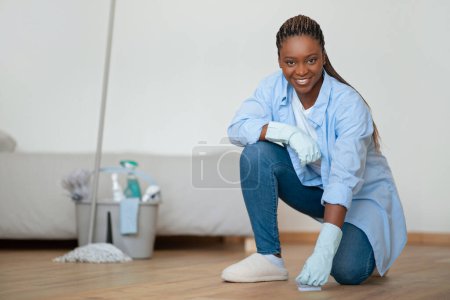 Téléchargez les photos : Happy attractive young african american woman in comfortable outwear housewife enjoying cleaning house, wearing rubber gloves, cleaning floor, smiling at camera, copy space. Chores, housekeeping - en image libre de droit