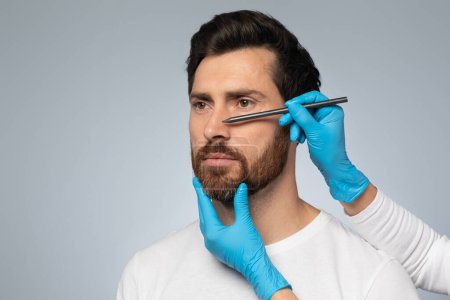 Téléchargez les photos : Portrait d'un homme d'âge moyen se préparant aux interventions de chirurgie plastique par un médecin dessinant des contours sur son nez, debout sur fond gris, espace de copie - en image libre de droit