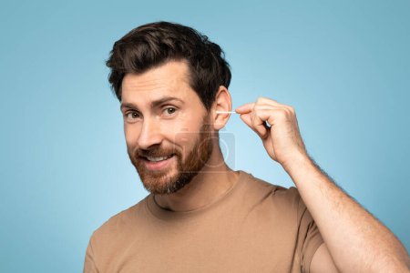 Téléchargez les photos : Happy handsome man using cotton swab stick for ear clean, posing over blue background and smiling at camera. Morning routine and skin care healthcare cosmetic procedures concept - en image libre de droit