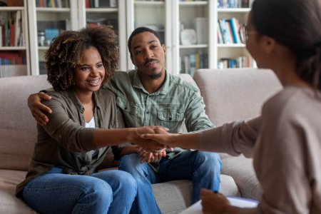 Photo for Happy beautiful young black couple in casual sitting on couch in cozy counselor office, embracing, shaking psychologist hand and smiling after successful marital family therapy, copy space - Royalty Free Image