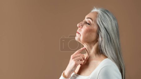 Foto de Calm mature european female with gray hair touches neck skin, enjoy treatment, isolated on brown background, studio, profile. Anti-aging cosmetics, beauty care, spa procedures result, ad and offer - Imagen libre de derechos