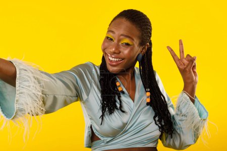 Téléchargez les photos : Blogger noir heureux femelle faisant Selfie geste signe de victoire, Posant ayant un maquillage lumineux debout sur fond jaune studio. Dame montrant V-signe sourire à la caméra - en image libre de droit