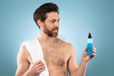 Téléchargez les photos : New beauty products. Handsome shirtless man holding bottle with blue cosmetic liquid, posing with towel on shoulder over blue studio background, free space - en image libre de droit