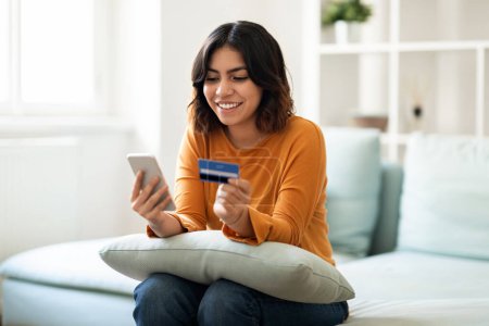 Téléchargez les photos : Purchasing Online. Smiling Arab Woman Using Smartphone And Credit Card At Home, Happy Young Middle Eastern Female Making Payments In Internet While Sitting On Couch In Living Room, Copy Space - en image libre de droit