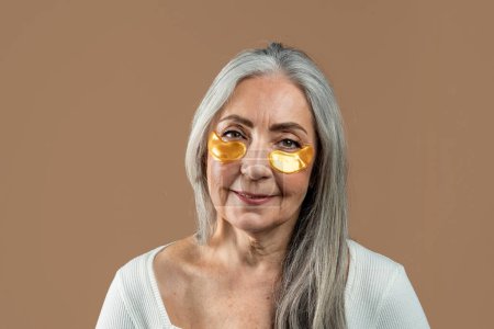 Téléchargez les photos : Cheerful old european lady with gray hair with gold patches under eyes, enjoys spa treatments, isolated on brown background, studio. Beauty care, anti-aging and anti-wrinkle procedures, ad and offer - en image libre de droit
