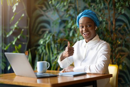 Photo for Positive happy attractive young black woman in casual and blue headscarf attending webinar, sitting at table, using modern laptop and earpods, showing thumb up at camera, cafe interior, copy space - Royalty Free Image