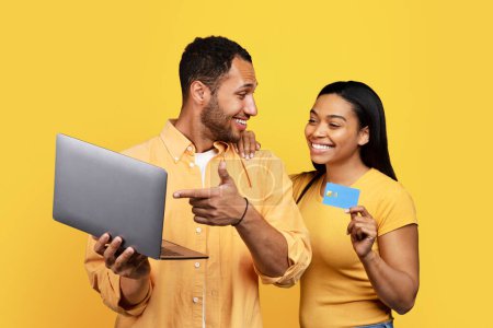 Téléchargez les photos : Glad young african american man pointing finger at laptop, female holding credit card, enjoy new buy, isolated on yellow background, studio. Cashback, online shopping purchases, finance and banking - en image libre de droit