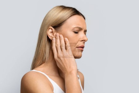 Tinnitus. Profil einer kranken blonden Frau mittleren Alters, die starke Ohrenschmerzen hat, ihren schmerzenden Kopf berührt, isoliert auf grauem Studiohintergrund, Panorama mit Kopierraum