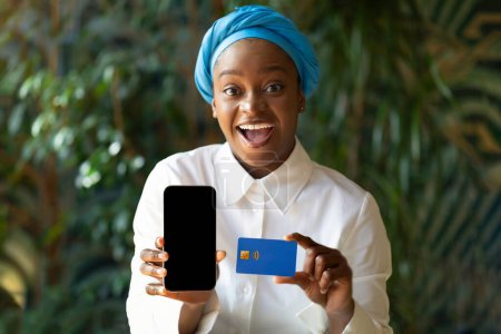 Téléchargez les photos : Excited happy attractive young black woman wearing headscarf showing cell phone with blank black screen and credit card and smiling, order bank card online, cafe interior, mockup, copy space - en image libre de droit