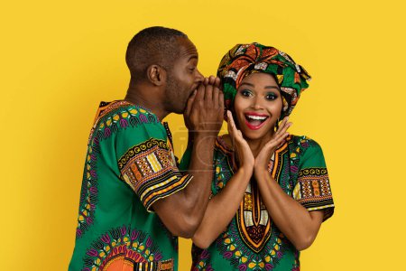 Foto de Handsome black man sharing secrets or rumors with his excited beautiful girlfriend, bright african couple in traditional costumes posing together on yellow studio background - Imagen libre de derechos
