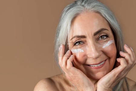 Téléchargez les photos : Cheerful senior european female with gray hair applies cream on face, enjoy daily procedure, isolated on brown background, studio. Skin and beauty care, anti-aging treatment, ad and offer, close up - en image libre de droit