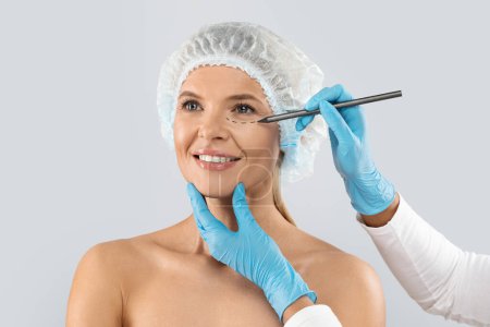 Foto de Plastic surgeon wearing blue medical gloves drawing contours with black pen around female patient eye before surgery, middle aged blonde woman in medical hat getting beauty treatment, isolated on grey - Imagen libre de derechos