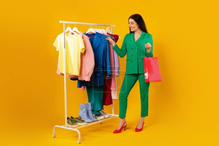 Téléchargez les photos : Happy female shopaholic standing with colorful shopper bags standing near clothing rail and choosing clothes over yellow background, full length. Lady buyer picking outfit - en image libre de droit