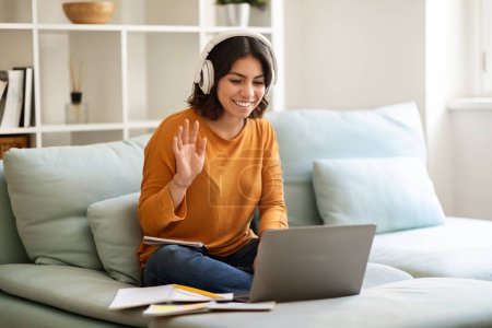 Téléchargez les photos : Jeune femme arabe heureuse dans les écouteurs faisant appel vidéo à la maison, souriante femme du Moyen-Orient utilisant un ordinateur portable pour la téléconférence, agitant à la caméra Web de l'ordinateur tout en étant assis sur le canapé, Copier l'espace - en image libre de droit
