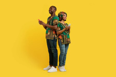 Photo for Mobile app. Beautiful cheerful happy loving black couple in colorful traditional african costumes standing back to back, using cell phones, posing over yellow studio background, copy space - Royalty Free Image