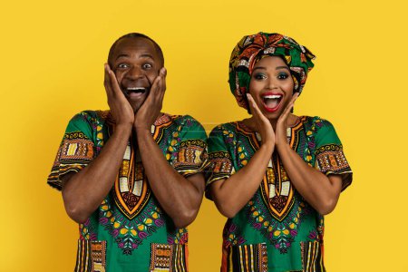 Photo for Excited loving attractive adult black man and woman in bright national african clothing posing on yellow studio background, looking at camera and touching face, showing amazement - Royalty Free Image