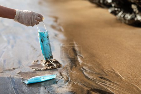 Téléchargez les photos : Femme main ramasser utilisé des masques médicaux jetables sur la plage tout en nettoyant la zone côtière, espace libre. Pollution plastique, déchets médicaux, ordures, déchets sur l'eau de mer - en image libre de droit