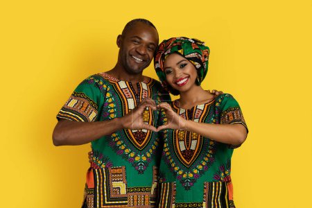 Foto de Beautiful cheerful black lovers middle aged man and millennial woman in traditional african costumes gesturing, showing heart-shaped symbol together, isolated on yellow studio background - Imagen libre de derechos