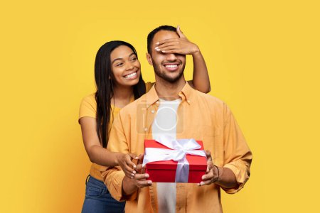 Photo for Cheerful young african american lady closes eyes with hand to guy, gives box with gift isolated on yellow background, studio. Birthday surprise, anniversary, holiday, Valentine day, celebrate Xmas - Royalty Free Image