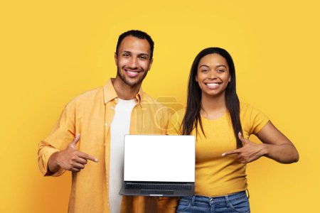 Photo for Smiling young african american husband and wife holding laptop with blank screen, recommend sale, app, ad and offer, isolated on yellow background, studio. Online shopping, blog and website, covid-19 - Royalty Free Image