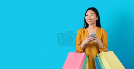Foto de E-commerce, online shopping, webstore concept. Happy attractive young korean woman with bunch of colorful shopping bags using smartphone on blue, looking at copy space and smiling, panorama - Imagen libre de derechos