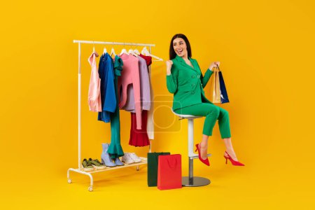 Téléchargez les photos : Concept d'achat et de vente. Femme shopaholic tenant des sacs shopper, assis près du rail de vêtements sur fond jaune, pleine longueur, espace libre - en image libre de droit