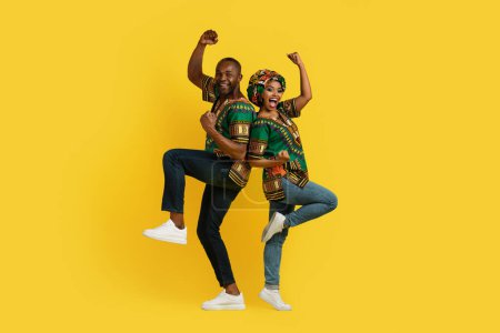 Téléchargez les photos : Positive happy cheerful black lovers man and woman in traditional african clothing celebrating success together on yellow studio background, jumping in the air and gesturing, full length - en image libre de droit