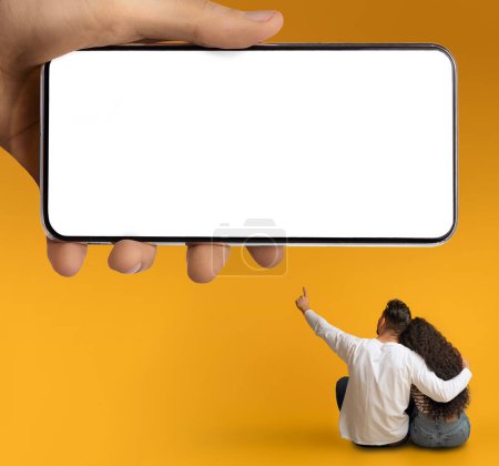 Photo for Couple Hugging Over Yellow Background And Pointing At Huge Blank Smartphone In Male Hand, Unrecognizable Man And Woman Demonstrating Free Place For Mobile Ad Or Design, Collage, Mockup - Royalty Free Image