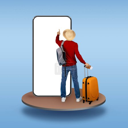 Foto de Elegante hombre con maleta naranja con sombrero de paja turista de pie en el pedestal sobre fondo de estudio azul, utilizando un gran teléfono con pantalla blanca en blanco, reserva de hotel o vuelo en línea, maqueta, collage - Imagen libre de derechos