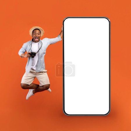 Téléchargez les photos : Full length of emotional African American man in summer clothes holding camera, jumping by huge phone with white screen mockup on orange studio background. Excited black guy going vacation, collage - en image libre de droit
