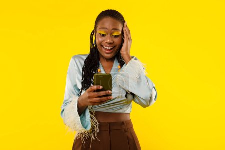 Téléchargez les photos : Femme afro-américaine excitée utilisant Smartphone SMS et lecture Wow offre mobile debout sur fond jaune Studio. Grande bannière publicitaire d'application mobile - en image libre de droit