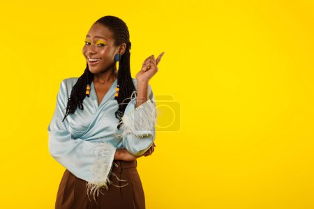 Photo for Cheerful Black Lady Pointing Finger Aside At Copy Space Advertising Great Offer Standing On Yellow Studio Background, Smiling Looking At Camera Posing With Bright Makeup. Advertisement Banner - Royalty Free Image
