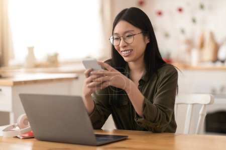 Photo for Cheerful happy pretty young korean woman in casual wearing eyeglasses entrepreneur working from home, using gadgets laptop and smartphone, checking newest business app, kitchen interior, copy space - Royalty Free Image