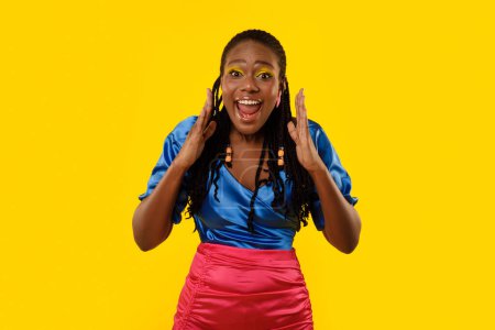 Téléchargez les photos : Cheerful African American Woman Shouting Hey Holding Hands Near Mouth Advertising Great Offer Standing On Yellow Studio Background. Lady Calling You Looking At Camera Posing With Bright Makeup - en image libre de droit