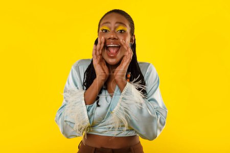 Photo for Hey. African American Female Shouting Holding Hands Near Opened Mouth Calling You And Advertising Great Offer Posing With Bright Makeup Over Yellow Studio Background, Looking At Camera - Royalty Free Image