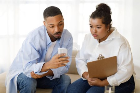 Foto de Handsome young black man patient going through difficult breakup or divorce, guy showing hispanic chubby woman psychologist shocking content on smartphone during therapy, counselor office - Imagen libre de derechos