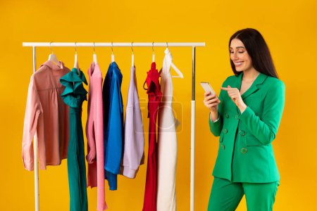 Photo for Shoppers application. Happy female customer using smartphone for purchasing new clothes online, standing near clothing rail on yellow background. Shopaholic woman with phone. - Royalty Free Image