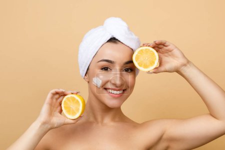 Téléchargez les photos : Skin Brightening. Smiling Young Indian Woman Holding Two Lemon Halves In Hands, Attractive Hindu Female With Towel On Head And Cream On Face Posing Over Beige Studio Background, Copy Space - en image libre de droit