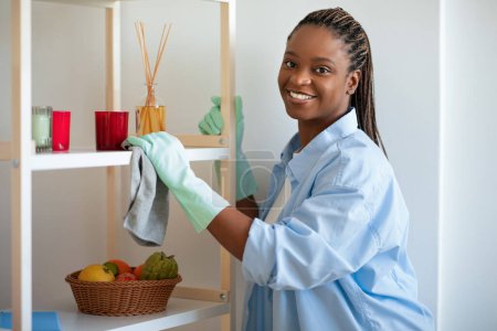 Photo for Happy attractive young black woman in comfortable outwear housewife enjoying cleaning house on weekend, wearing rubber gloves, cleaning dust, smiling at camera, copy space. Chores, housekeeping - Royalty Free Image