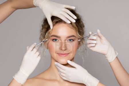 Photo for Attractive young woman getting injection for eyes area by two cosmetologists, beautician hands in gloves giving anti aging serum shot on female face against grey background - Royalty Free Image