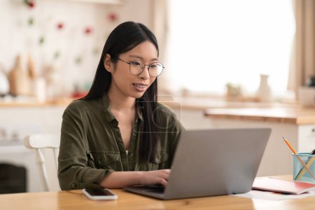 Photo for Happy pretty millennial asian lady in casual wearing eyeglasses freelancer or entrepreneur working from home, sitting at kitchen table, using modern laptop, typing on keyboard, copy space - Royalty Free Image