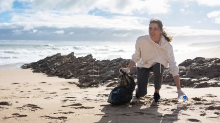 Foto de Earth day. Female volunteer with polyethielene bag picking up plastic bottle on the beach, cleanup garbage on the ocean coast, panorama with free space. The concept of conservation of ecology - Imagen libre de derechos