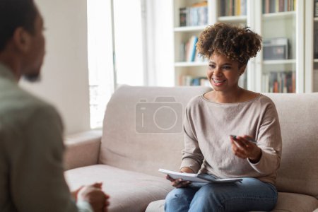 Photo for Psychological help, support. Smiling friendly pretty young black woman in casual psychologist sitting on couch, holding folder, having conversation with patient african american man, copy space - Royalty Free Image