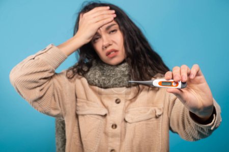 Téléchargez les photos : Sad millennial european female in scarf presses hand to forehead, suffers from headache and fever, show thermometer isolated on blue background, studio. Ill, flu and cold, health problems, covid-19 - en image libre de droit