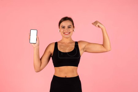 Photo for Fitness App. Sporty Young Woman Holding Smartphone With Blank Screen And Showing Arm Muscles, Athletic Female Demonstrating Empty Cellphone While Standing Isolated Over Pink Background, Mockup - Royalty Free Image